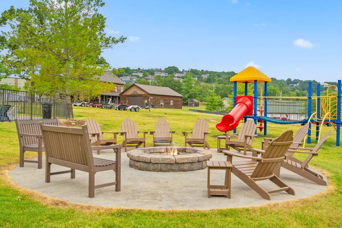 This exterior shot captures a cozy fire pit surrounded by Adirondack chairs, a playground with equipment for children, and lush green surroundings. The scene evokes relaxed evenings with families gathered around the fire, children playing, and a sense of community bonding under the stars.