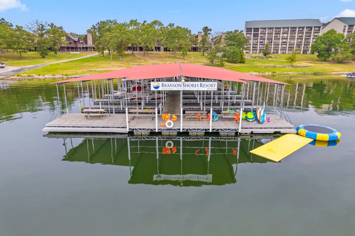 A vibrant exterior shot of a dock extending over the tranquil lake, with floating mats, life rings, kayaks, and shimmering water inviting guests to participate in swimming, fishing, boating, or simply relaxing by the water’s edge. The image radiates a sense of fun and recreation, with equipment and landscape that suggest all-ages enjoyment and direct access to lake activities.