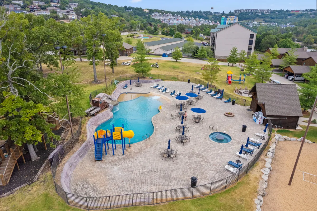 This exterior shot showcases the sparkling swimming pool with a beach entry, colorful playground equipment, hot tub, lounge chairs, and a sand volleyball court, all surrounded by lush landscaping. People can be seen swimming, sunbathing, and socializing, capturing a lively, resort-like environment.