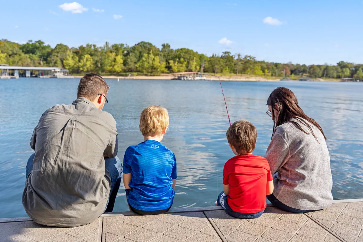A family is pictured sitting together by the water, each holding fishing rods, as they enjoy quiet bonding time on the shore. The calm water and relaxed posture of the family members evoke the peaceful and communal moments described in the narrative.