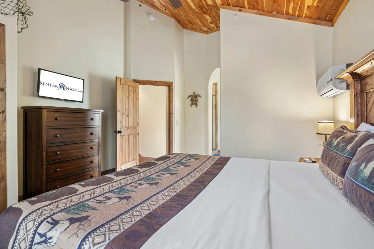 This image features a cozy bedroom with a comfortable bed, a wooden chest of drawers, a wall-mounted TV, and an arched doorway. The room exudes an inviting atmosphere ideal for relaxation, with soft lighting and touches that suggest both comfort and privacy.