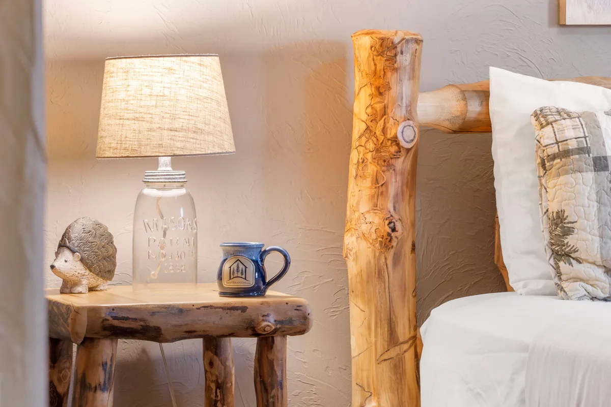 A cozy bedside scene with a rustic wooden lamp, hedgehog figurine, plaid pillow, blue mug, and basket. Natural light filters in, enhancing the inviting feel of the bedding and decor.