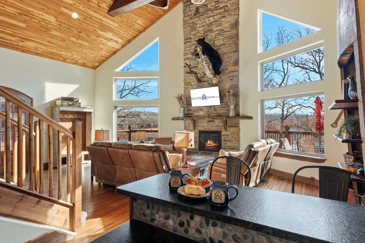 A cozy seating area arranged before a large stone fireplace, with sunlight streaming through oversized windows. The wooden staircase and rustic decor add charm, while comfortable sofas invite groups to relax, converse, or celebrate.