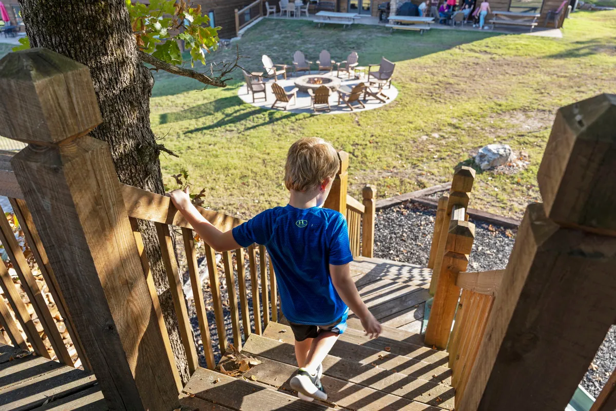 A cozy outdoor fire pit area with a young boy descending wooden stairs, surrounded by seating on a wooden deck. The atmosphere is relaxed and friendly, perfect for evening gatherings, roasting marshmallows, and enjoying crisp fall weather.