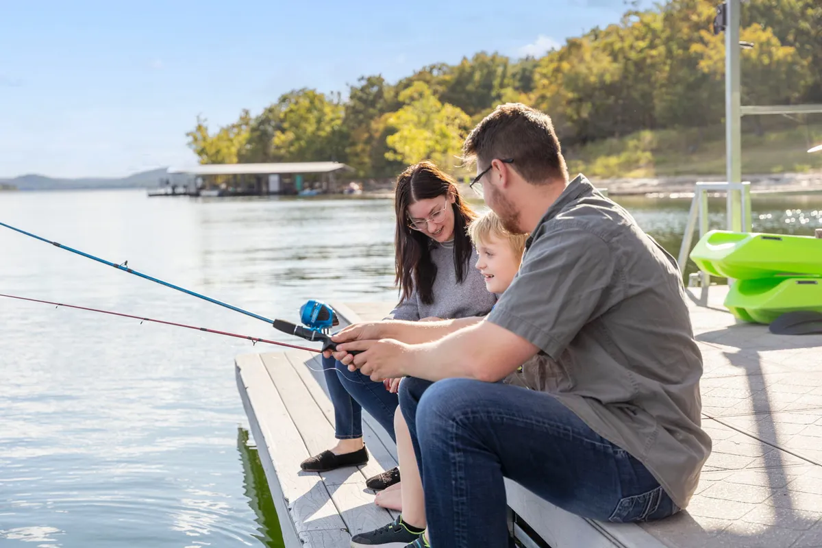 A joyful lakeside moment showing family members fishing from the dock, with the calm lake in the background. The dock and water are clear, emphasizing both the accessibility of boating and fishing, and the natural beauty of the setting.