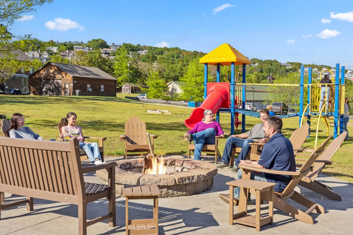 An inviting scene featuring a fire pit with people gathered around sharing stories, adjacent to playground equipment and a comfortable seating area. Children are visible playing nearby, and the sunset creates a warm, communal ambiance.