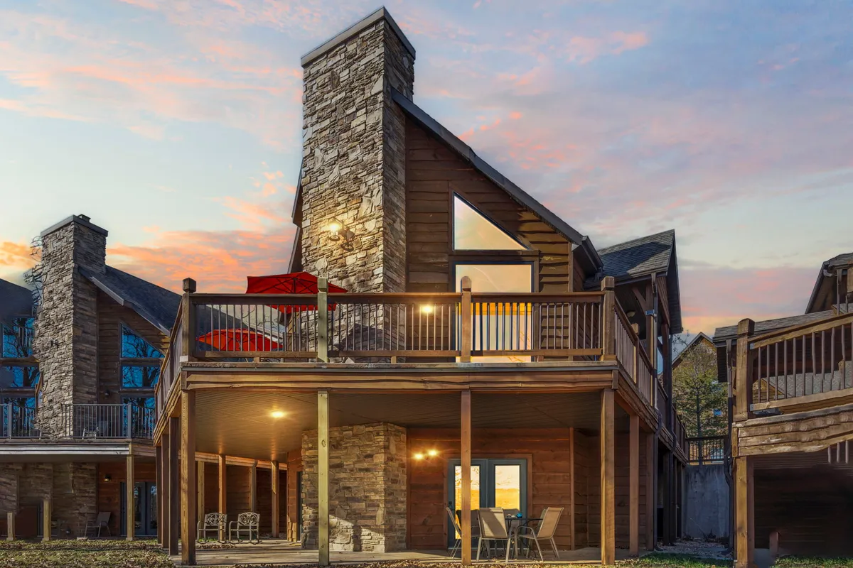 An expansive deck with vibrant red umbrellas, modern glass doors leading into the lodge, and a large stone chimney. Outdoor seating areas offer views of the lake and provide space for dining and evening gatherings under the open sky.