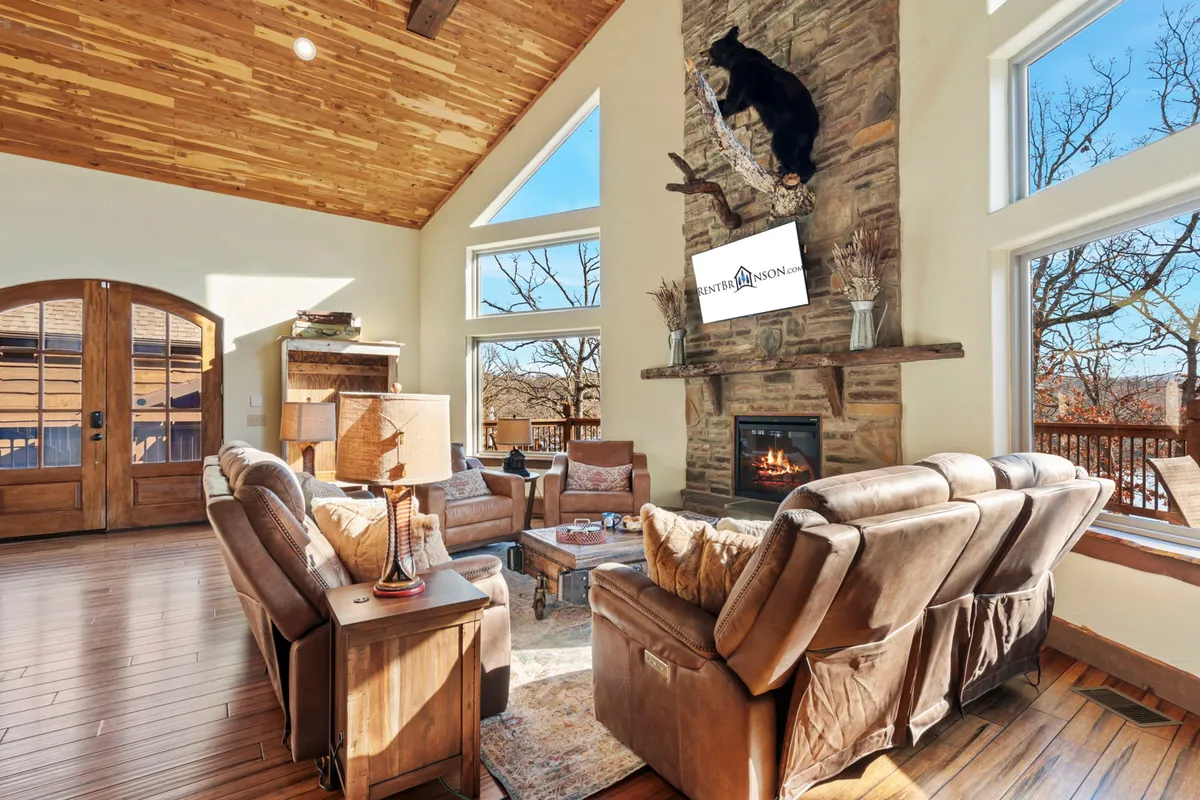 The image features a warm, inviting living room with a grand stone fireplace as the centerpiece, flanked by a mounted bear for a rustic touch. Large windows fill the space with natural light and frame scenic lake views, while plush seating beckons guests to relax and connect.