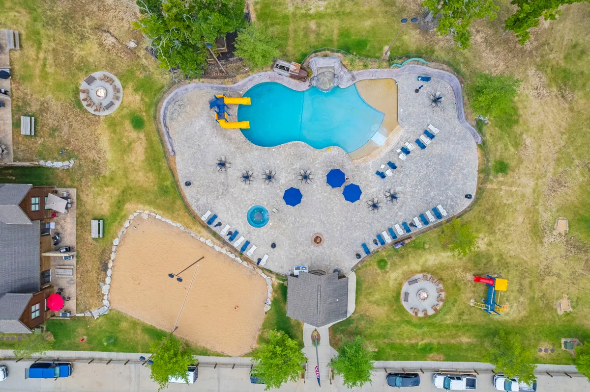 This image showcases a vibrant outdoor scene with a swimming pool featuring a water slide, a beach volleyball court, a hot tub, and lounge chairs. The setting is lively with people enjoying the various amenities, from splashing in the pool and sliding down the water slide to playing volleyball and relaxing in the hot tub. In the background, the fire pit area suggests social gatherings as the sun begins to set.